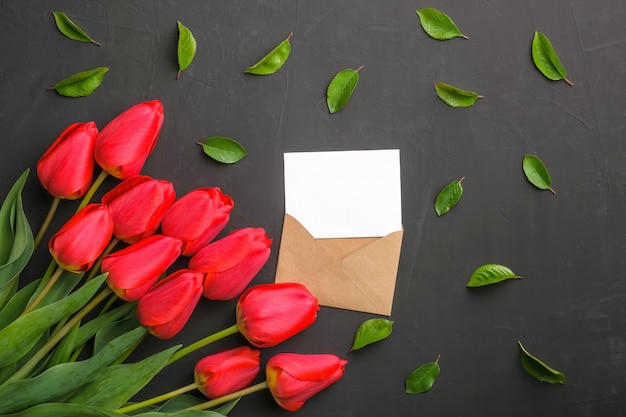 Mockup of fresh red tulips bouquet and greeting card in kraft envelope and scattered leaves