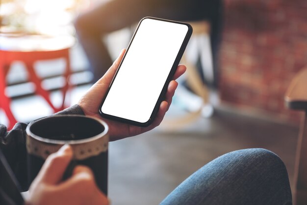 Mockup image of a hand holding black mobile phone with blank screen while drinking coffee in cafe