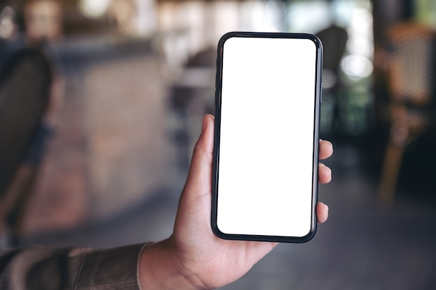 Mockup image of a hand holding and showing black mobile phone with blank white screen in modern cafe