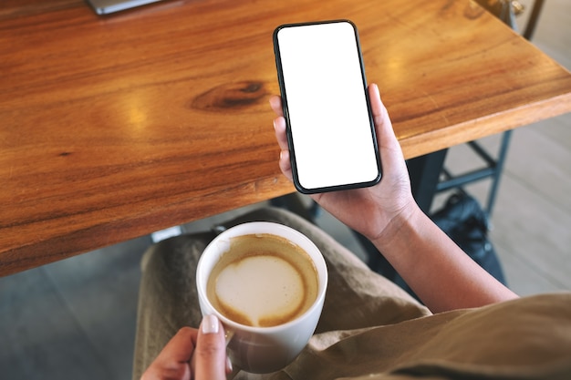 Mockup image of a woman holding black mobile phone with blank desktop screen while drinking coffee