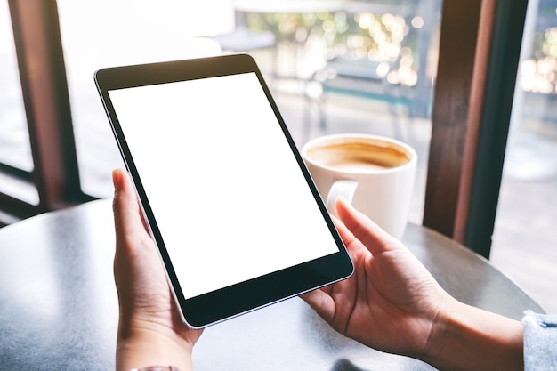 Mockup image of a woman holding black tablet with white blank screen and coffee cup on the table