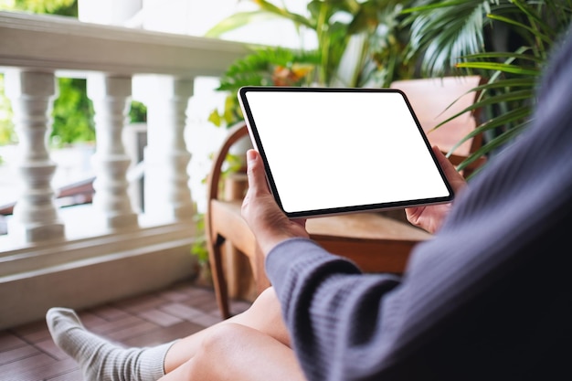 Mockup image of a woman holding and using digital tablet with blank white desktop screen while sitting on balcony at home