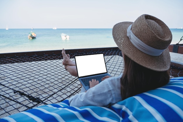 Mockup image of a woman using and typing on laptop computer with blank desktop screen while lying down on hammock on the beach