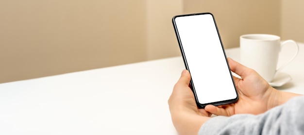 Mockup mobile cell phone hands of woman with cell phone against background of yellow wall and cup of coffee female hand holding using mobile on desk banner