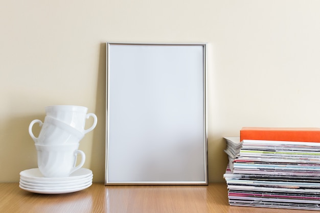 Mockup template with large A4 silver frame, stack of magazines and ceramic cups with plates on wooden table next to yellow wall.