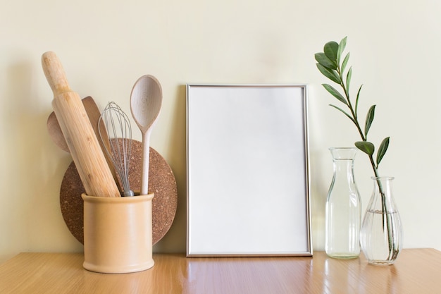 Mockup template with large A4 silver frame, wooden kitchen utensils and Zamioculcas plant in glass vase.