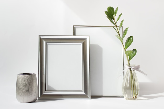 Mockup template with two blank silver frames, silver small vase and green Zamioculcas plant branch in glass vase.