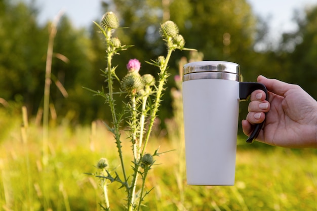 Mockup of a woman holding a white travel mug by a burdock flower Empty mug mock up for promotion