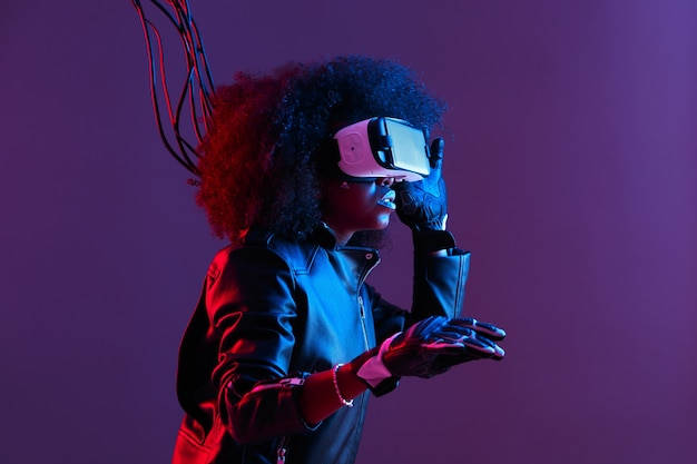 Mod curly dark haired girl dressed in black leather jacket and gloves uses the virtual reality glasses on her head in the dark studio with neon light . Cyberpunk character