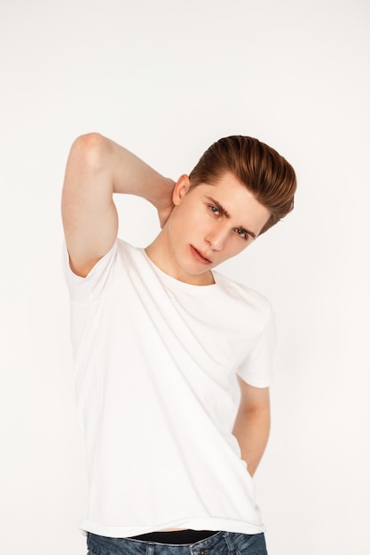 Model European young fashionable man with stylish hairstyle in trendy youth casual clothes from new summer collection stands near vintage wall in studio. Fresh portrait pretty handsome guy indoors.