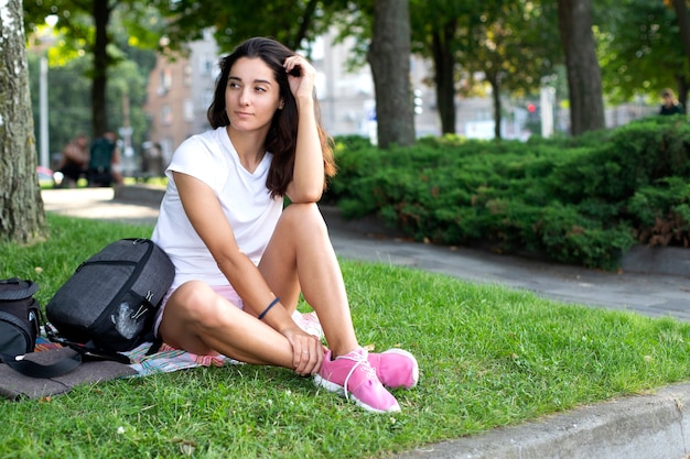 Model girl brnetka in a white shirt and sneakers, sits on the grass in the park. Blurred background, place for an inscription.