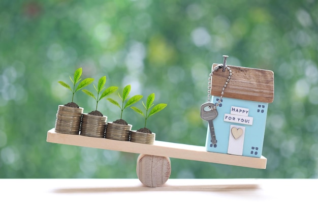 Model house with Trees growing on stack of coins money on wood scale seesaw