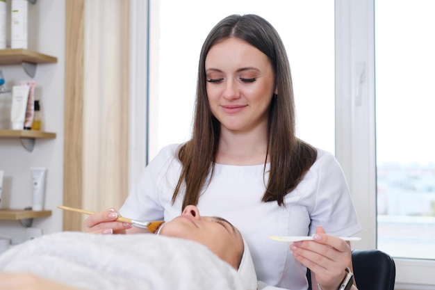 The model lies on the couch with her eyes closed Cosmetologist in blue gloves touching the patient's face with a brush Cosmetic clinic Health care clinic cosmetology