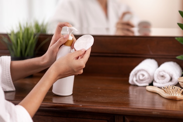 A model in a white bathrobe squeezes a product from a white bottle, which stands on a brown wooden dressing table, on which towels, a comb and a flower pot stand, on a cotton pad. Close-up.