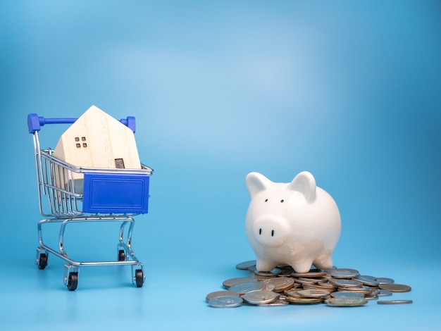 A model wooden house on shopping cart With a pile of coins and Piggy bank on blue background.