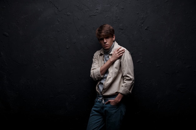 Model young man in a light stylish spring jacket in a trendy knitted gray golf with a fashionable haircut in vintage blue jeans posing indoors near the black wall. American style men's clothing