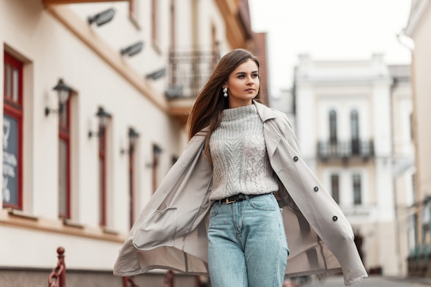 model of a young woman in a trench coat and a knitted vintage sweater walks down the street