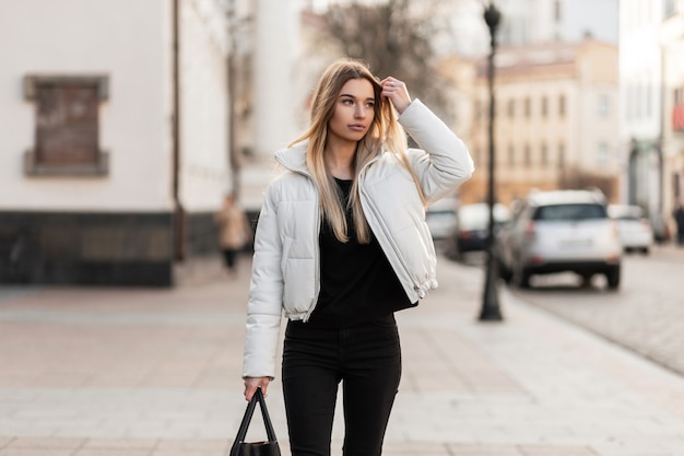 model of a young woman in a white jacket and a leather trendy bag stands on the street