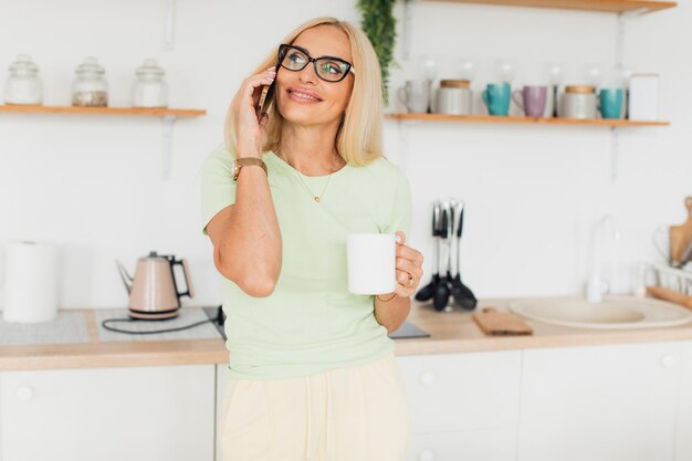 Modern attractive middle-aged woman talking on the phone and drinking coffee in the kitchen at home