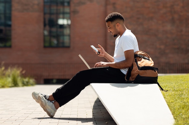 Modern black man with laptop and cellphone sitting outdoors and surfing internet or texting side