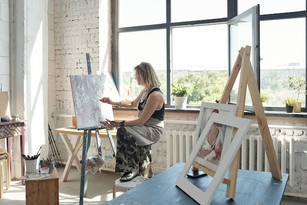 Modern blond-haired artist in glasses holding foot on stool and painting on canvas using palette