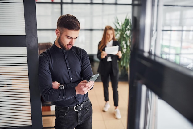 Modern businessman standing indoors in office. His female colleague behind.