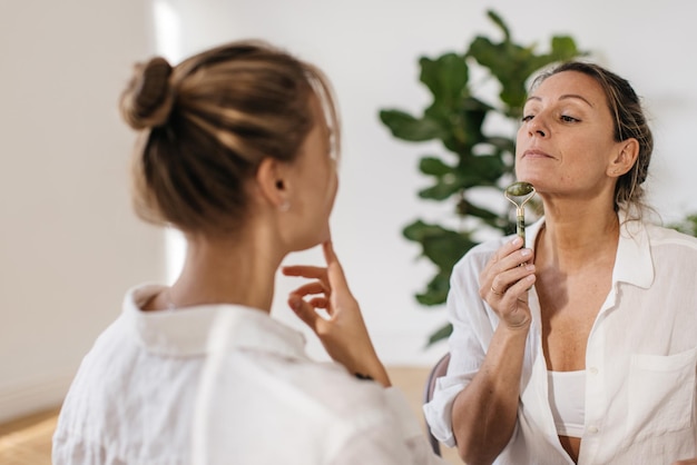 Modern caucasian ladies in white shirts doing face massage with quartz roller indoors. Wellness and self care concept