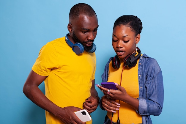 Modern couple using smartphone and headset to browse internet, standing over blue background. Happy man and woman enjoying social media app on mobile phone with touch screen in studio.