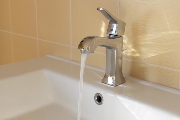 Modern faucet in yellow bathroom with running water, close up, side view. Concept hygiene, house cleaning, water saving, supply problems, reducing use, conscious consumption. Horizontal.