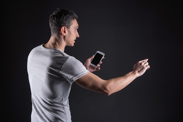 Modern gadget. Nice handsome young man holding his smartphone and looking at the computer monitor while touching it