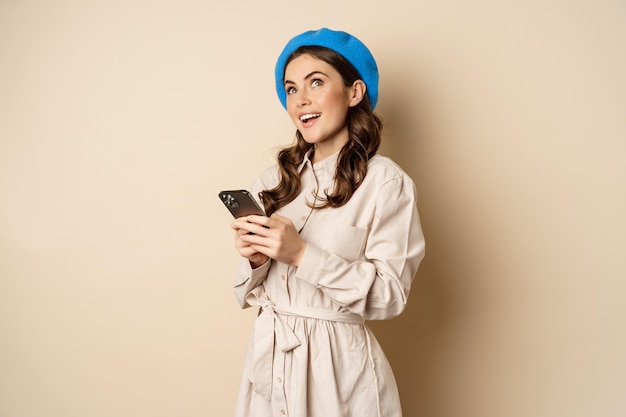 Modern girl using smartphone smiling and posing happy with mobile phone standing over beige background