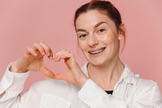 Photo modern happy woman smiling with braces and showing heart with hands on pink background