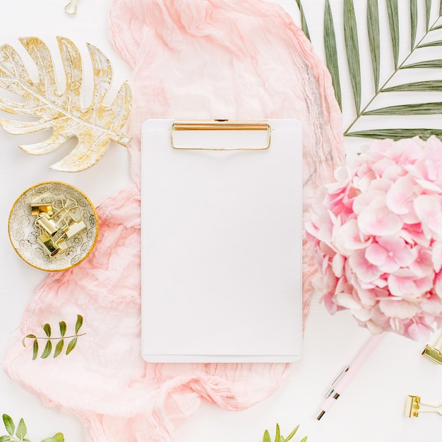 Modern home office desk workspace with blank paper clipboard, pink hydrangea flowers and accessories on white surface