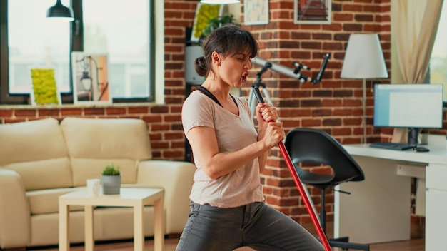 Modern housewife acting funny mopping living room floors, sweeping dirt and dust with mop and having fun. Young adult smiling, listening to music and dancing at home, cleaning apartment.