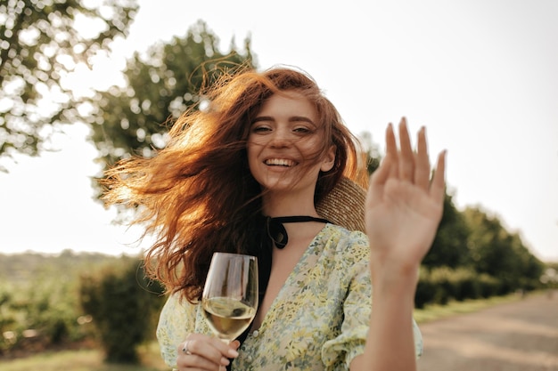 Modern lady with red long hair in green and yellow printed dress laughing looking into camera and holding glass with wine outdoor