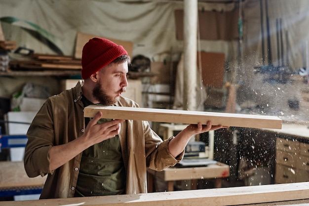 Modern male carpenter wearing red knit cap holding wood plank blowing dust from it in joinery workshop