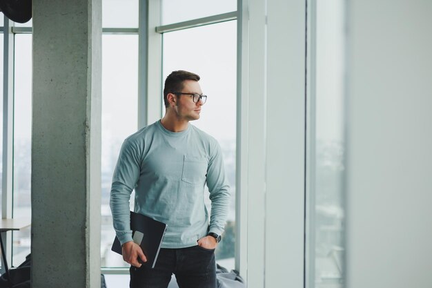 Modern male manager in casual clothes stand by the window in the workspace with a laptop Male freelancer works remotely