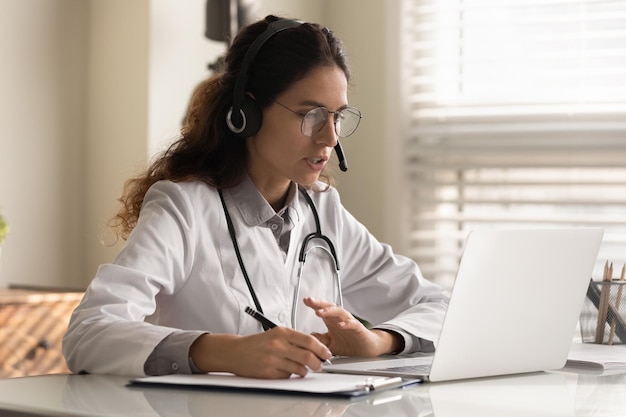 Modern medicine Focused young female doctor general practitioner in headset watching medical webinar video conference on laptop screen taking notes Young woman medic intern learning online using pc