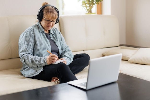 Modern middle aged blond woman in headphones take online course or training on computer at home Smart mature Caucasian female in earphones watch webinar make note study distant on laptop