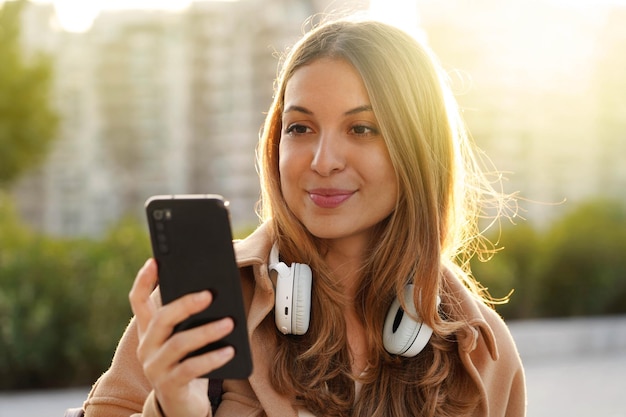 Modern millennial chooses a song from her playlist on smartphone to listening with headphones outdoors at sunset