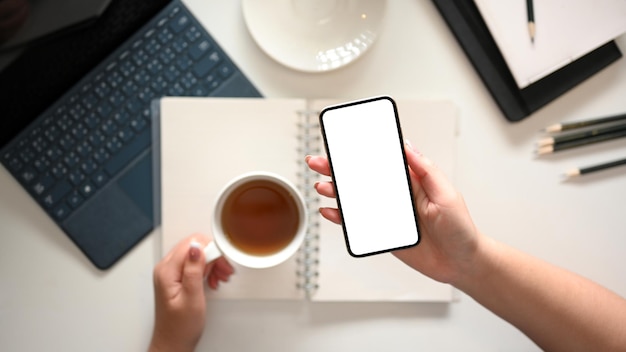 A modern minimal white workspace with a female hands holding a teacup and smartphone mockup