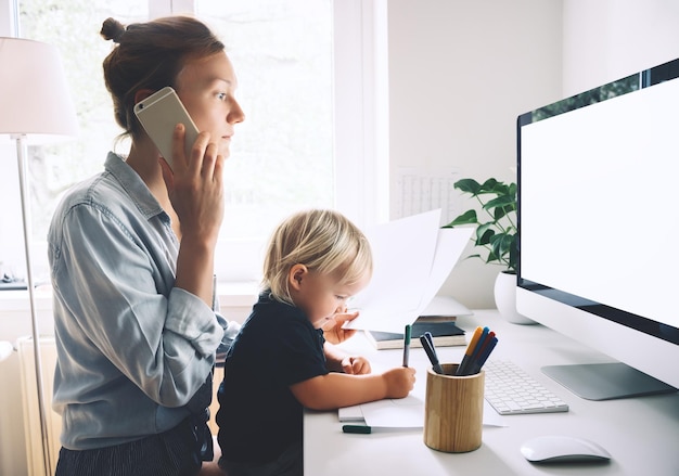 Modern mom balances between work and child home schooling on sick leave or quarantine Mother works from home with kid Woman responding on phone calls working on computer with a toddler on her lap