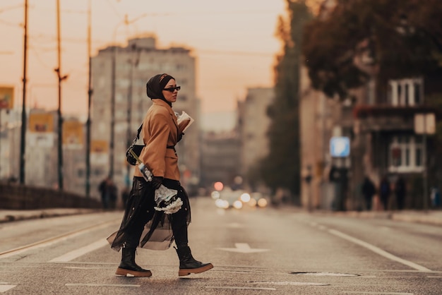 A modern Muslim woman with a hijab walks around the city while carrying a newspaper and bread in her hand.