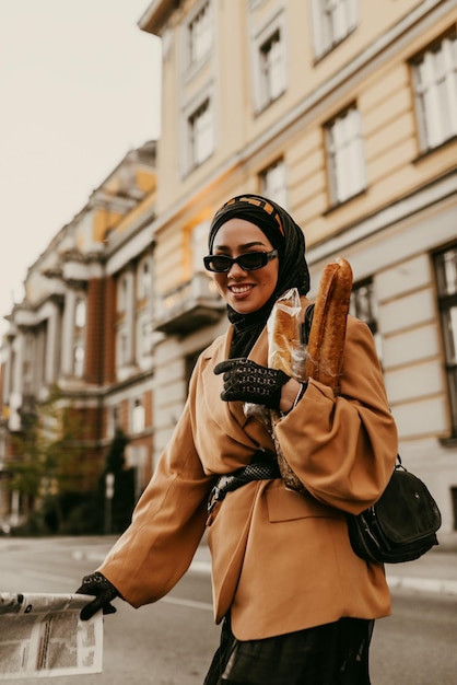 A modern Muslim woman with a hijab walks around the city while carrying a newspaper and bread in her hand.