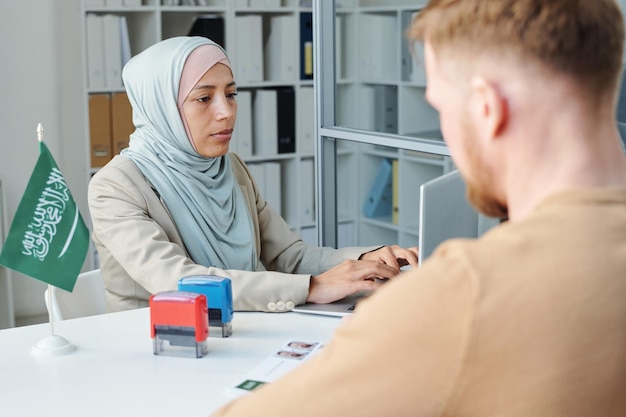 Modern muslim woman working in saudi arabia embassy consulting unrecognizable man for visa application