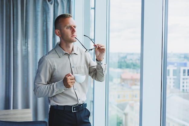 Modern office businessman or manager drinking coffee by the window smiling Portrait of successful IT engineer programmer wearing glasses working in office