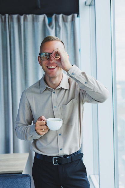 Modern office businessman or manager drinking coffee by the window smiling Portrait of successful IT engineer programmer wearing glasses working in office