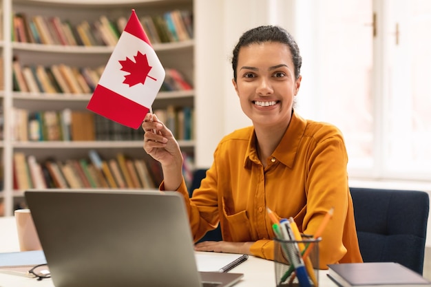 Modern online foreign education Happy mixed race woman holding flag of Canada using laptop sitting in library