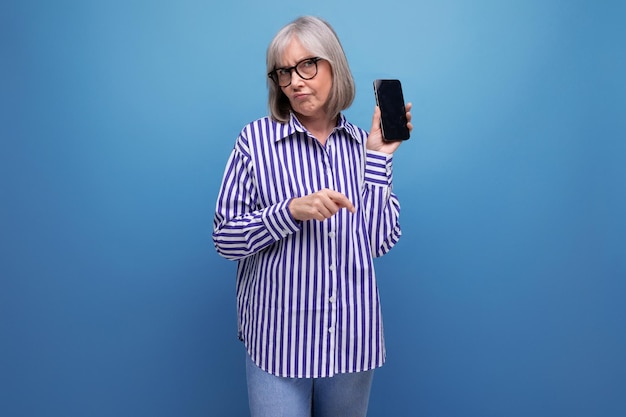 Modern pensioner middleaged woman with gray hair is studying smartphone gadget on bright studio