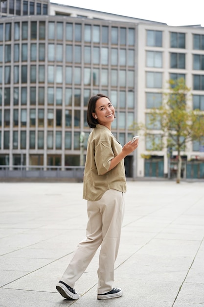 Modern people young woman walks on street of city and listens music in wireless headphones holds sma